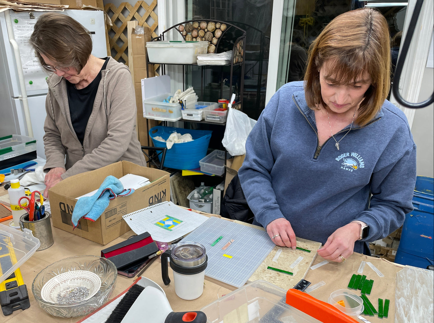 Two women working at a table with stained glass items including a box, tools, and papers.