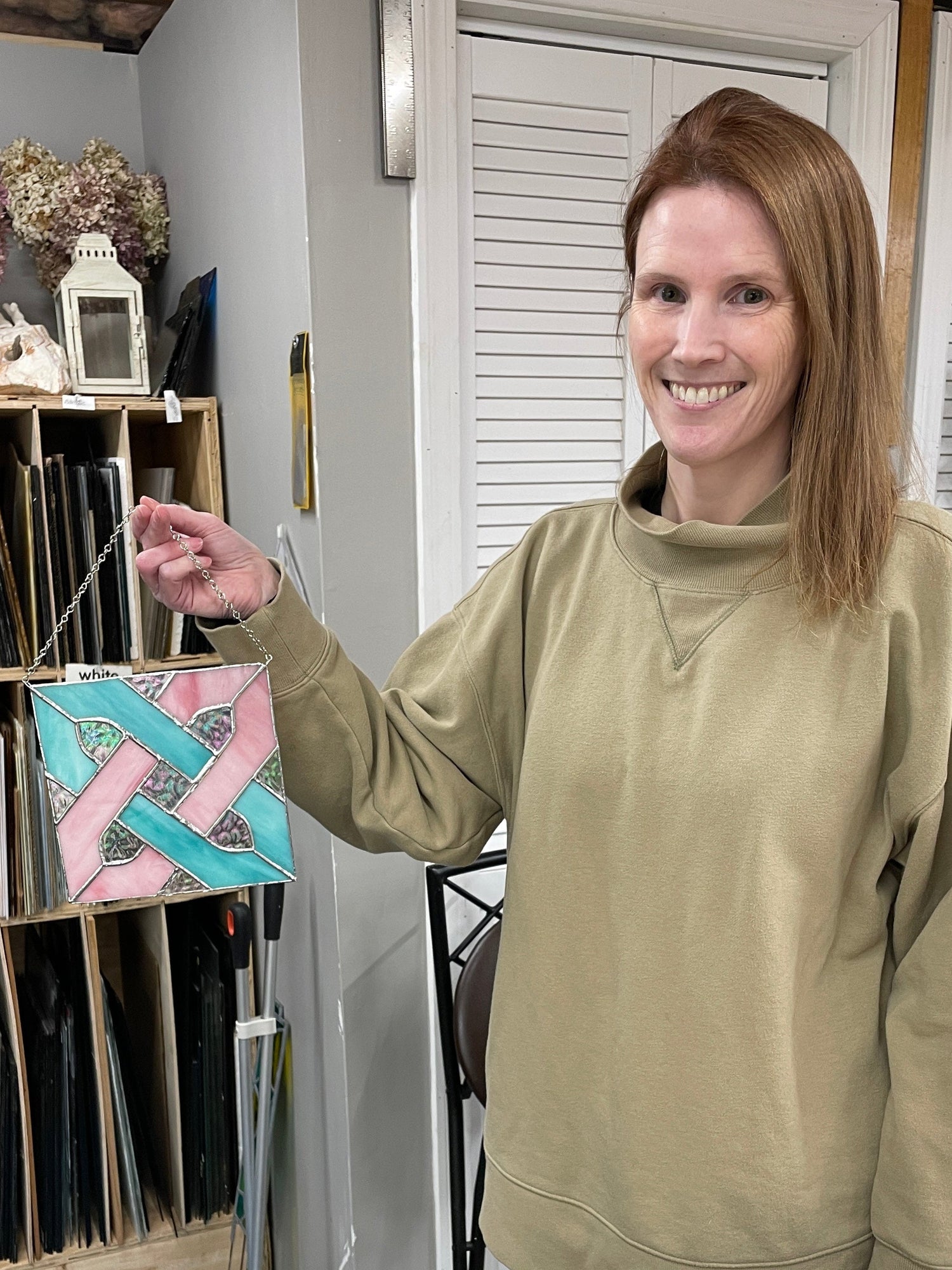 Smiling stained glass student holding a finished pink and teal celtic knot panel she made in a beginner class
