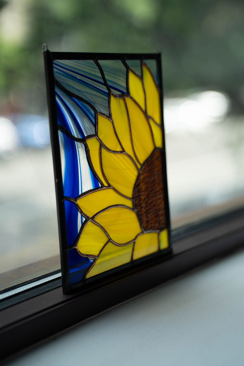 Stained glass sunflower panel on a window sill with a blurred outdoor background