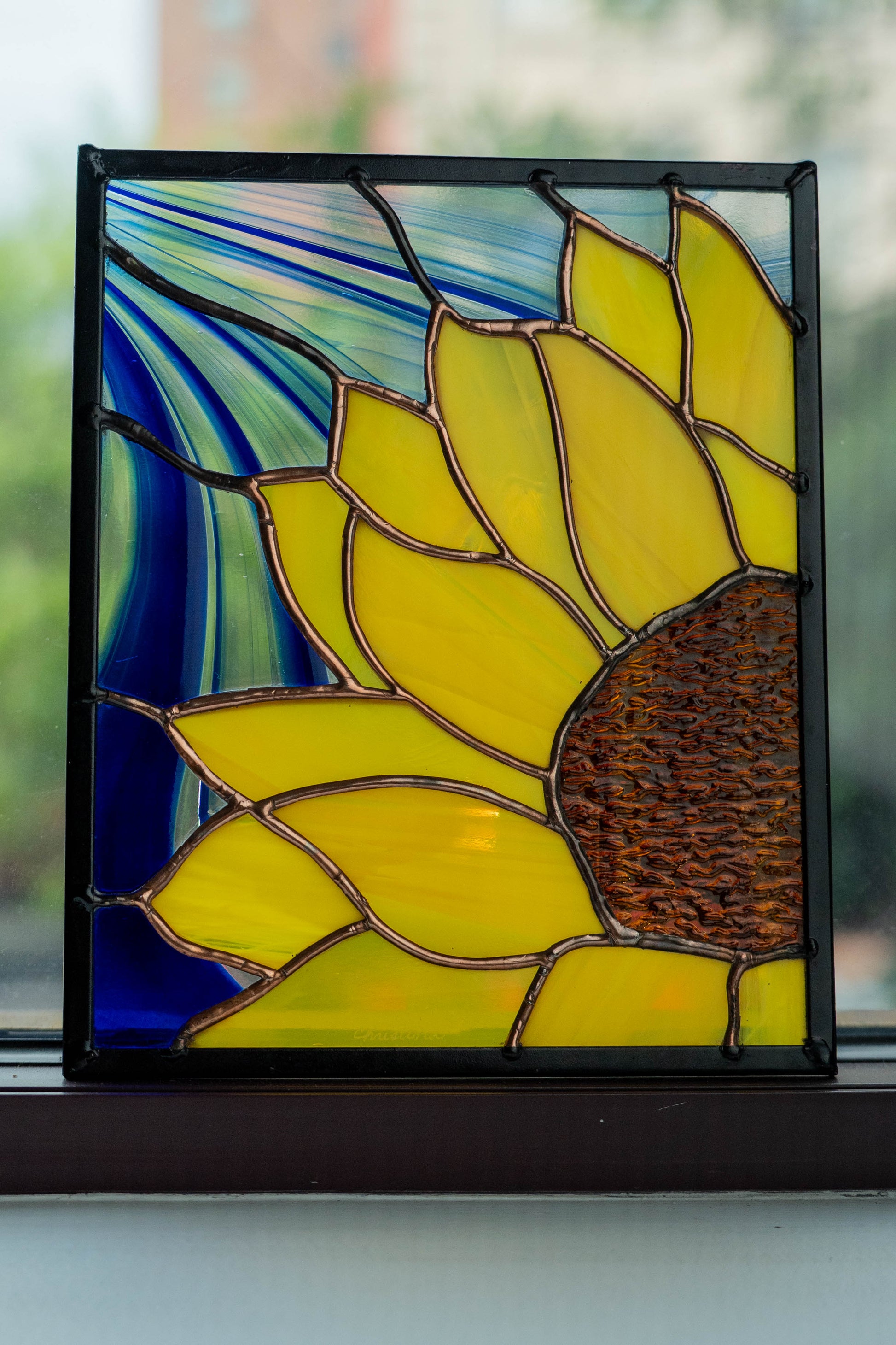 Stained glass sunflower panel on a window sill with blurred greenery in the background